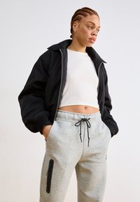 Veste bombardier noire avec des poignets côtelés, assortie à un pantalon de survêtement gris avec des poches zippées sur les côtés et une taille élastiquée avec cordon.