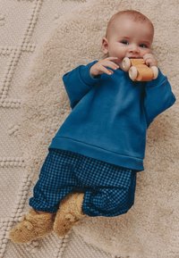 Modra jopa, modre in črno karo hlače ter rjavi puhasti copati na otroku, ki drži leseno igračo in leži na teksturiranem svetlem preprogi.