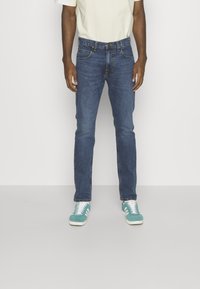 Hombre vestido con jeans ajustados azules, camiseta blanca y zapatillas turquesa, de pie frente a un fondo blanco liso.