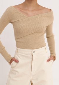 Žena ve béžovém žebrovaném svetru s odhalenými rameny, dlouhými rukávy a krémových kalhotách s vysokým pasem, ruce v kapsách, neutrální pozadí.