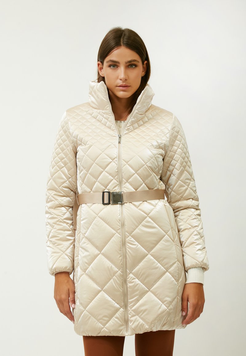 Abrigo acolchado tipo puffer en beige claro, con un cuello alto, cremallera de longitud completa y una cintura ceñida con un cinturón y una hebilla metálica. Diseño texturizado.