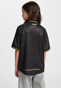 Svart kortärmad tröja med geometrisk textur, gula detaljer på kragen och ärmarna, samt "INTERNATIONALS" tryckt på baksidan.