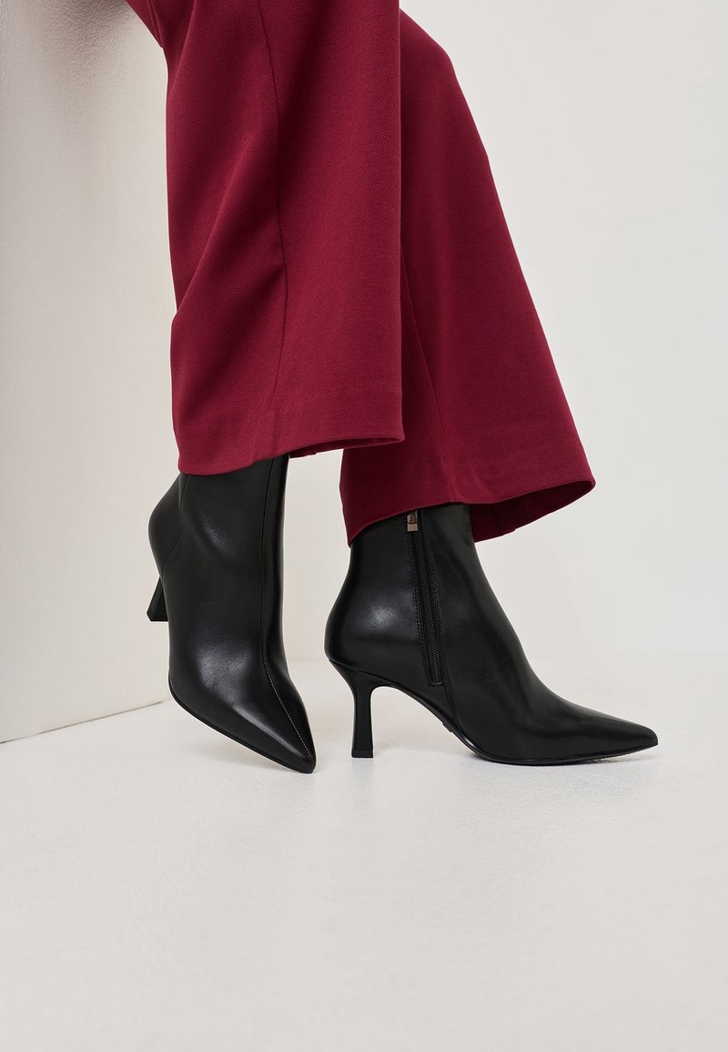 Bottines noires à bout pointu avec fermeture éclair latérale et talons fins portées avec un pantalon large rouge foncé sur fond uni.