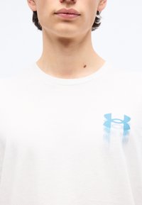 Noor, kes kannab valget särki, millel on rindkerel tuhm sinine Under Armouri logo ja kaelas väike sünnimärk.