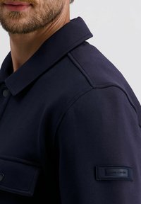 Marineblauwe jas met hoge kraag, zijzakken en een gestructureerde stof. Heeft een label op de mouw met de tekst "Pierre Cardin."