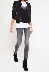 Chaqueta bomber de satén negra sobre una blusa negra con detalles de encaje, combinada con jeans skinny grises y botines negros.