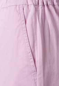 Lichtroze stof met een lichte glans, met gestikte details en een zakopening. Het materiaal lijkt zacht en ademend te zijn.