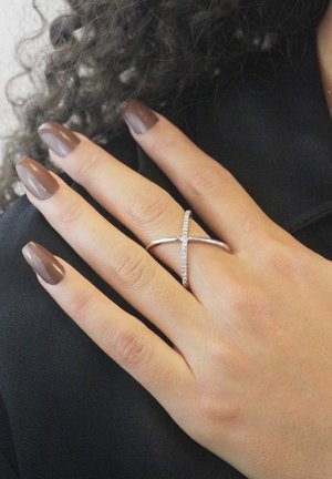 Silberner Ring mit einem Kreuzmuster, verziert mit kleinen klaren Steinen, getragen an einer Hand mit braunen Nägeln, vor einem dunklen Hintergrund.