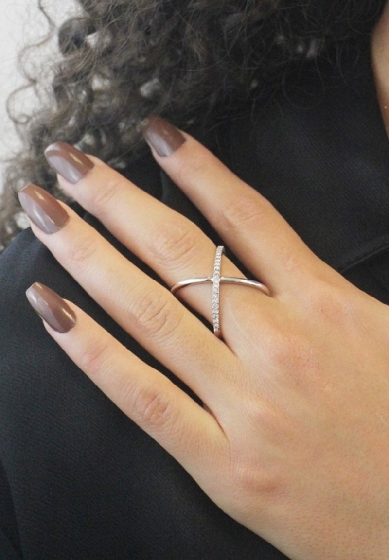 Anello d'argento con un design incrociato, decorato con piccole pietre trasparenti, indossato su una mano con unghie marroni, su uno sfondo scuro.