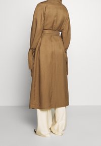 Długi brązowy płaszcz trench w talii, wykonany z gładkiego materiału, z długimi rękawami z wiązanymi detalami, zestawiony z szerokimi kremowymi spodniami.