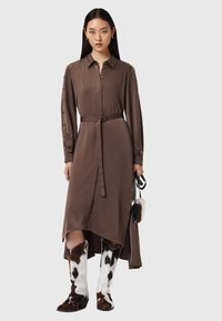 Abito di raso marrone con maniche bottonate, cintura e orlo asimmetrico. Abbinato a stivali al ginocchio con stampa maculata e una piccola borsa.