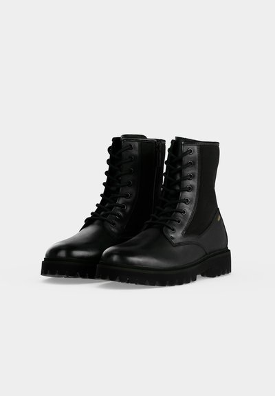 Paire de bottes de combat en cuir noir avec semelles épaisses, laçage à l'avant et fermetures éclair latérales sur un fond clair et uni.