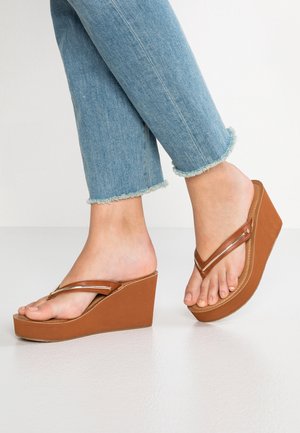 Fødder iført brune kile-sandaler med brede stropper og lyseblå jeans med filtreret kant, stående på en lys gulv mod en hvid baggrund.