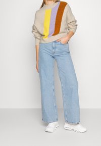 Femme portant un pull beige avec des rayures jaunes, violettes et brunes, un jean large bleu clair, des baskets blanches, debout avec la main dans la poche.