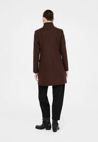 Cappotto marrone con un design strutturato, colletto alto e vestibilità su misura. Indossato su pantaloni neri e stivali alla caviglia, che mettono in risalto una texture liscia.