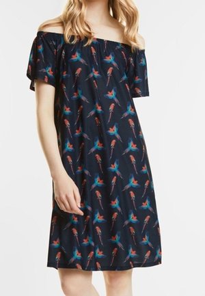 Kvinde iført en marineblå off-shoulder kjole med et farverigt fuglemønster, stående med den ene arm ned langs siden mod en ensartet hvid baggrund.