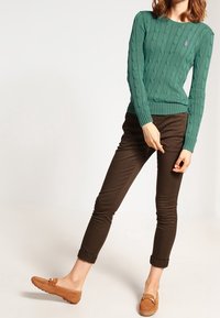 Grön kabelstickad tröja med ribbad nederkant, ihop med bruna slim-fit byxor och beige moccasin-liknande skor; diskret logotyp accent på tröjan.