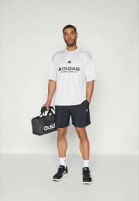 Vyras, vilkintis baltą Adidas sportinę marškinėlį, juodas šortus, juodus sportinius batelius, baltas kojines, laikantis juodą Adidas sportinę kuprinę, stovi prieš paprastą foną.