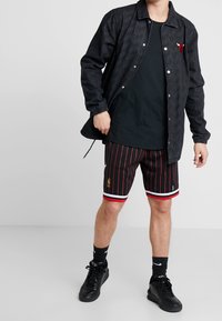 Mann som bär en svart Chicago Bulls-jacka, svart t-shirt, svarta och röda randiga NBA-shorts, svarta Nike-strumpor och svarta sneakers.