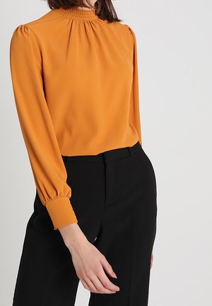Mujer con blusa de manga larga color mostaza con puños fruncidos y pantalones negros de cintura alta, de pie frente a un fondo liso.
