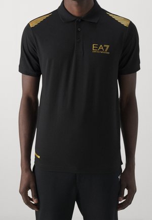 Zwarte poloshirt met korte mouwen en gouden EA7 Emporio Armani-logo op de borst, met gouden strepen op de schouders, gedragen door een staande persoon.