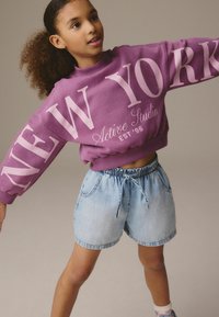Felpa viola con grande testo bianco "NEW YORK" e "Active Studio EST '96". Abbinata a pantaloni shorts in denim blu con vita elastica.