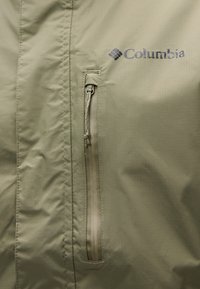 Khakifarbene Columbia-Jacke mit Reißverschlusstasche, glattem Stoff mit leichtem Glanz und gesteppten Details. Logo deutlich sichtbar.