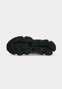 Semelle noire de basket avec motifs de grip texturés et dessin de bande de roulement segmenté, montrée allongée à plat sur une surface gris clair.