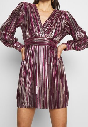 Donna che indossa un vestito lungo a maniche lunghe a righe viola e argento lucido con scollatura a V profonda e vita stretta, in piedi con le mani sui fianchi.