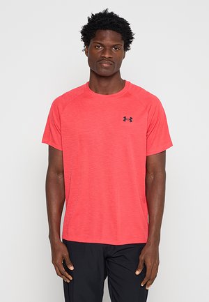 Mężczyzna z krótkimi kręconymi włosami, ubrany w jasnoróżową koszulkę z krótkim rękawem Under Armour i czarne spodnie, stoi na jednolitym tle.