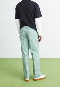 Lichtgroene broek met rechte pijpen gemaakt van katoen met een gladde textuur, voorzien van achterzakken en een merkpatch. Draagt erbij een zwart shirt.