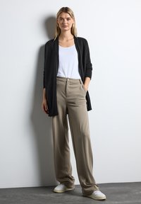 Czarny kardigan zapinany z przodu na białym t-shircie, w połączeniu z beżowymi spodniami o szerokich nogawkach i białymi trampkami na szarym podłożu.