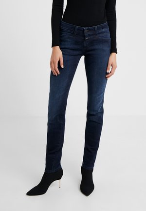 Femme portant un jean skinny bleu foncé, un haut noir à manches longues et des bottines chaussettes noires à talons, debout devant un fond blanc uni.