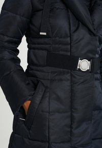 Svart puffjacka med kviltad textur, som har ett brett bälte med ett silverfärgat sp�änne och sidofickor. Varm och isolerad design.