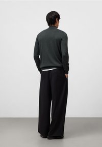 Maglione lavorato a maglia verde scuro con colletto alto, abbinato a pantaloni neri ampi. La texture è morbida, offrendo una vestibilità comoda.