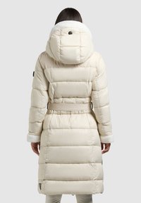 Steppmantel in Hellbeige mit tailliertem Schnitt, gefütterter Kapuze mit Fleece und gerippten Bündchen. Glatte, gesteppte Textur mit klar abgegrenzten Bereichen.