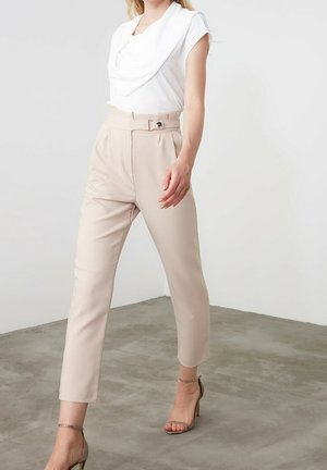 Femme portant un haut blanc drapé, un pantalon beige taille haute avec ceinture, et des sandales à talons hauts beige à lanières, debout sur un sol en béton.