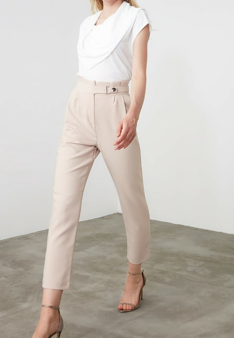 Femme portant un haut blanc drapé, un pantalon beige taille haute avec ceinture, et des sandales à talons hauts beige à lanières, debout sur un sol en béton.