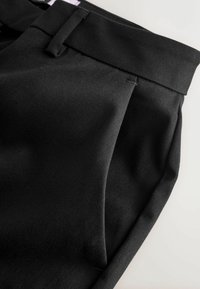 Pantalon noir avec une finition lisse et texturée, doté d'une ceinture plate, de poches latérales et de coutures ajustées.