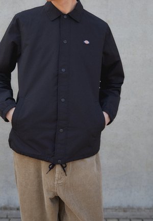 Personne portant une veste noire boutonnée avec un logo sur la poitrine et un pantalon en velours côtelé beige, les mains dans les poches, debout contre un mur clair uni.