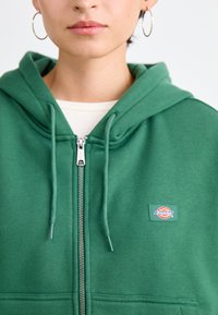 Green zip-up hoodie made of soft fabric, featuring a logo patch on the chest, metal zipper, and drawstring hood.