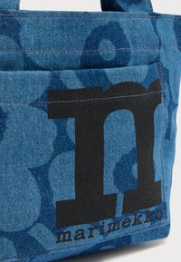 Τσάντα ώμου από μπλε τζιν με μεγάλο μαύρο «m» και λουλουδάτα μοτίβα, με το όνομα της μάρκας «marimekko» τυπωμένο στην κάτω δεξιά γωνία.