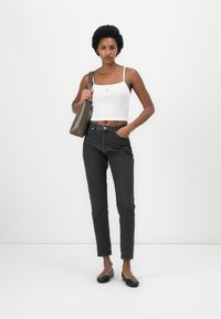 Top corto bianco a coste con spalline regolabili, abbinato a jeans neri in denim a vita alta. Tiene in mano una borsa in pelle verde oliva con texture.