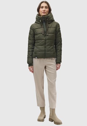 Olijfgroene pufferjack met capuchon, ritssluiting en koordjes, gecombineerd met lichtbeige broek en beige enkelboots.