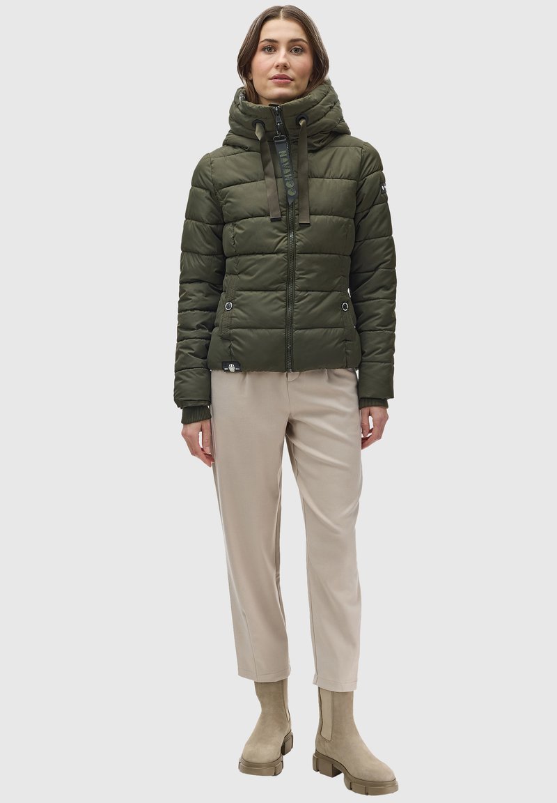 Chaqueta puffer verde olivo con capucha, cierre de cremallera y cordones, combinada con pantalones beige claros y botas de tobillo beige.