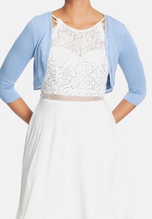 Femme portant une robe en dentelle blanche avec un shrug bleu sans manches, mains posées sur les hanches, sur fond uni.