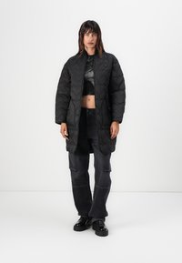 Zwarte gewatteerde lange jas met een staande kraag, met een gladde textuur en zijzakken, gedragen over een crop top en baggy donkere jeans.
