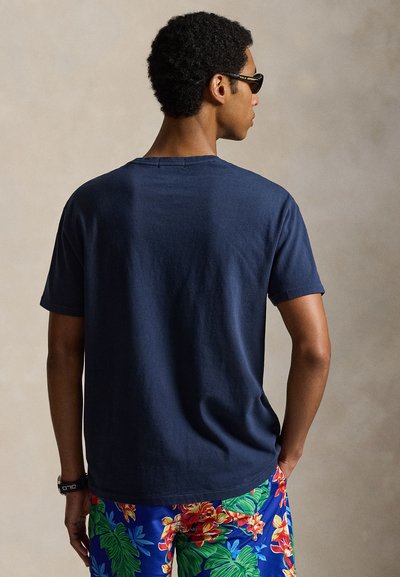 Άνδρας που φοράει μαύρα γυαλιά ηλίου, ναυτικό μπλε μπλουζάκι και μπλε floral σορτς, στέκεται με την πλάτη προς ουδέτερο φόντο.