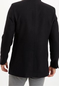 Blazer nero in lana con silhouette aderente. Presenta cuciture sartoriali e due bottoni sui polsini, visto da dietro.