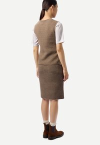 Femme portant un gilet brun en tricot sans manches et une jupe assortie jusqu'aux genoux, une chemise blanche à manches courtes, des chaussettes beiges et des bottines marron.